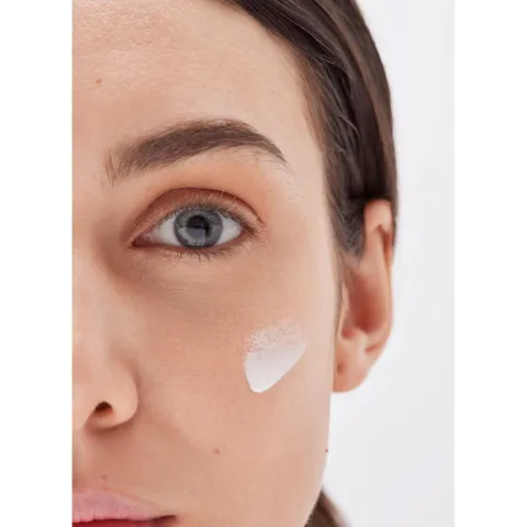 Woman with fair skin and blue eyes applying cream to her cheek.