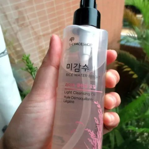 Hand holding a clear bottle of Korean cleansing oil with a black pump, greenery in background.