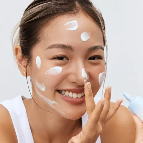 Smiling woman applying white cream to her face, wearing a white top.