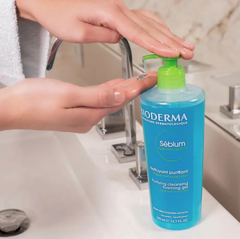 Hands using a blue Bioderma cleanser bottle with a green pump at a bathroom sink.