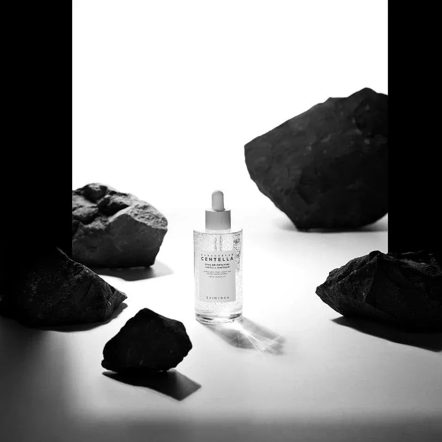 Clear dropper bottle of serum surrounded by dark rocks on a white background.